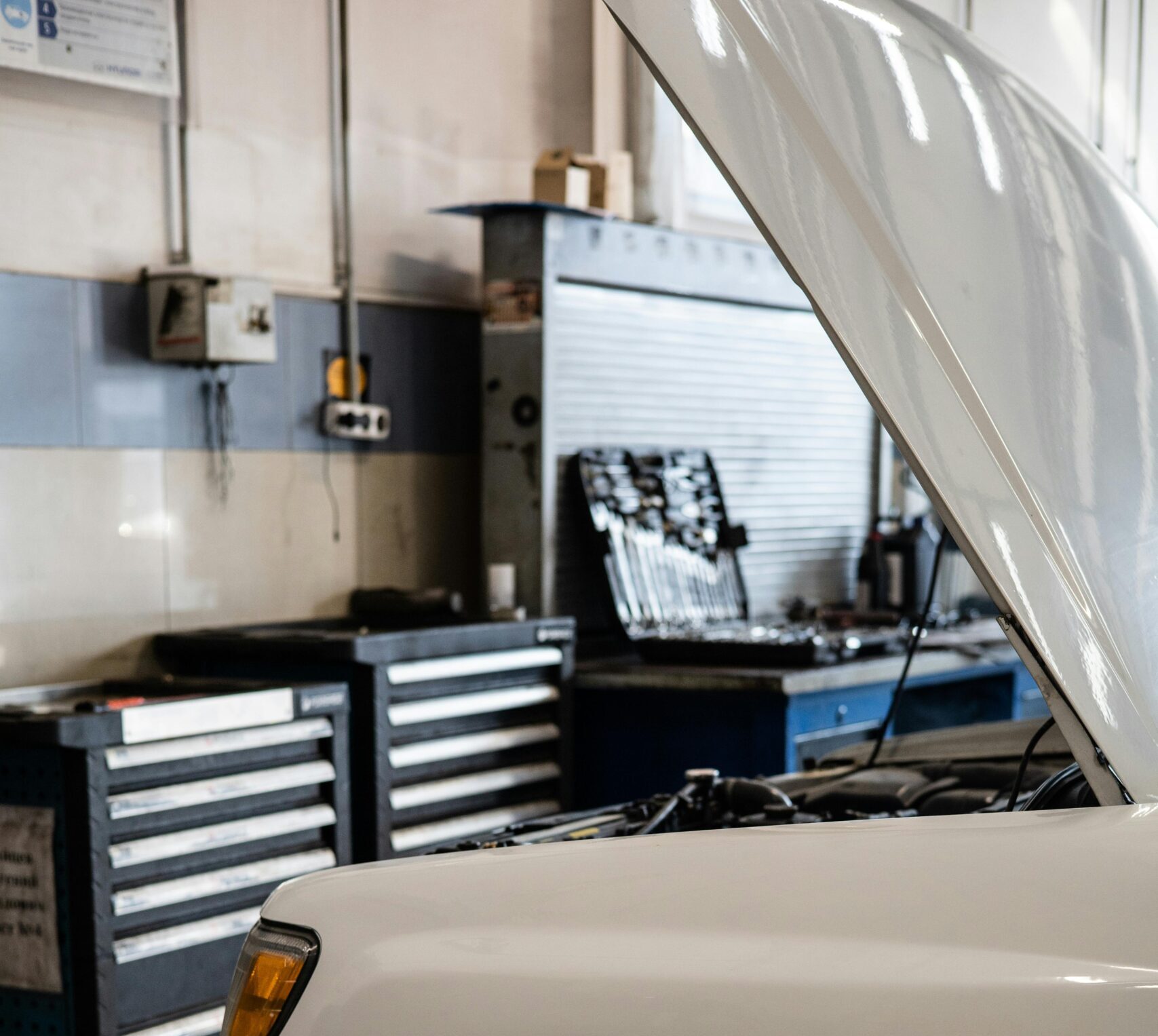 White car with hood open in a professional auto repair shop. Toolbox and equipment visible.